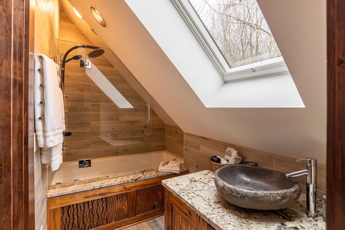 A spa-like bathroom with a deep, jetted tub set below a skylight, a sculpted stone sink, and elegant finishes. The space is flooded with natural light, suggesting the luxury of soaking while gazing at the sky—whether it’s clouds by day or stars by night.