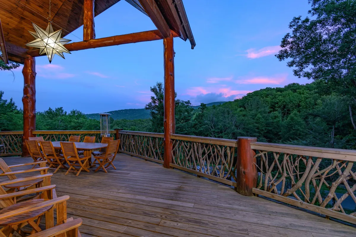 A spacious wooden deck surrounded by lush trees features a large dining table, star-shaped hanging light, and a decorative railing. The scene is set for outdoor dining and relaxed gatherings, with the ambiance suggesting both daytime and evening enjoyment.