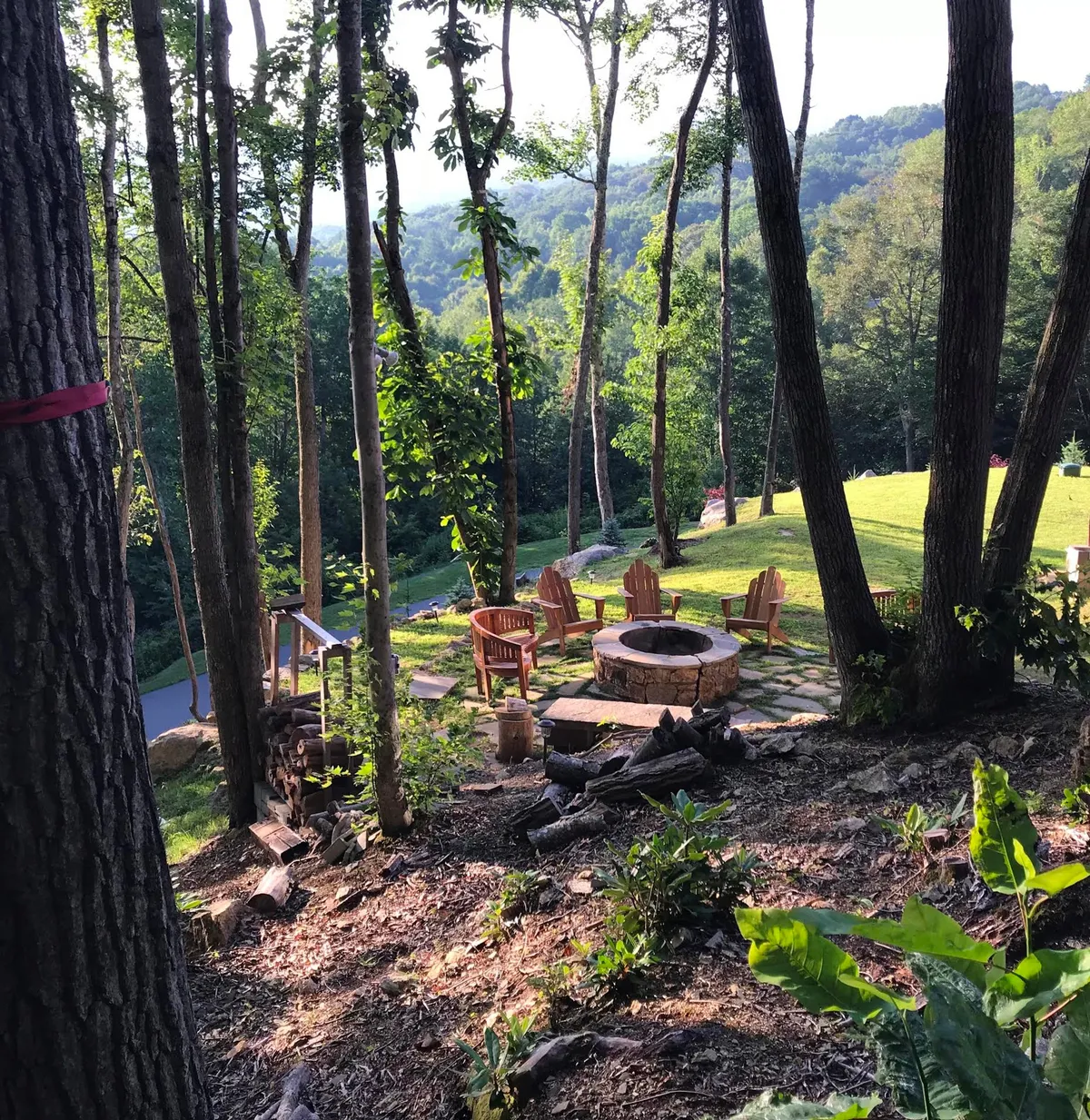 A welcoming fire pit surrounded by Adirondack chairs sits amidst a lush, tree-lined landscape. The meandering pathway hints at opportunities for hiking or peaceful strolls, with the setting sun bathing the area in warm light.