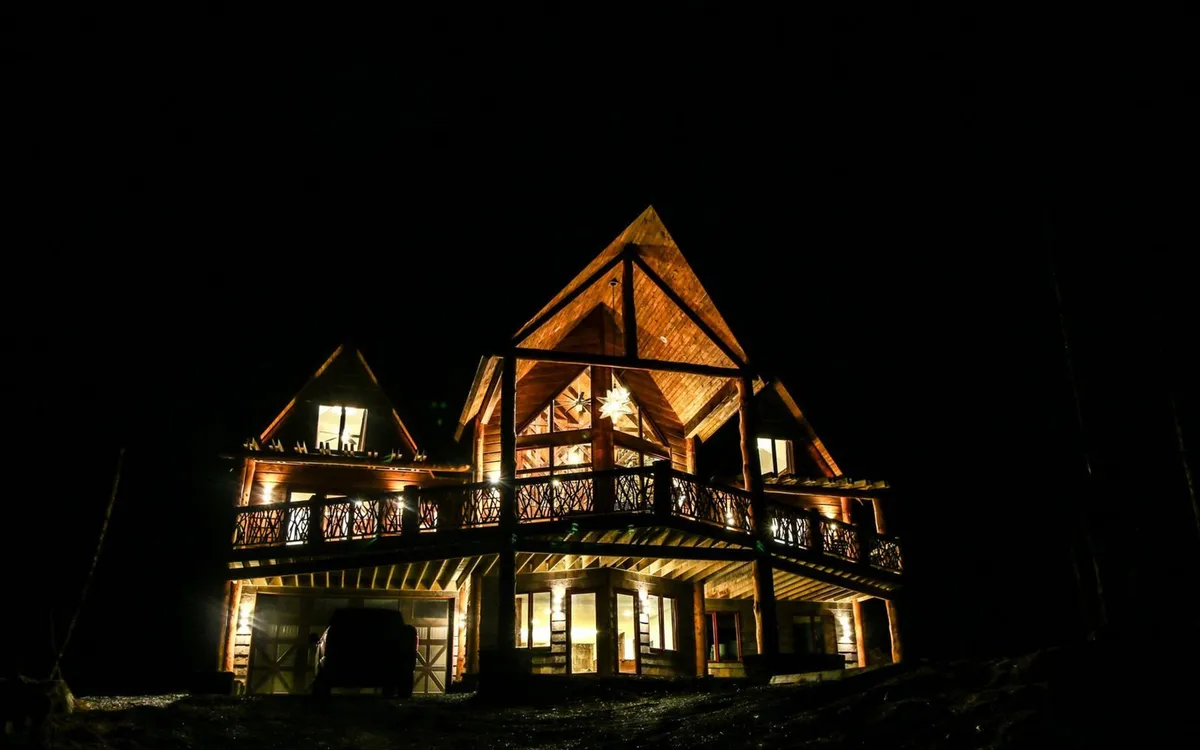 This image features the illuminated facade of the Copperleaf home at dusk, highlighting its intricate wooden architecture and inviting balcony with decorative railing. The warm glow from the interior and exterior lighting contrasts beautifully with the surrounding natural landscape, giving a strong sense of both privacy and grandeur.