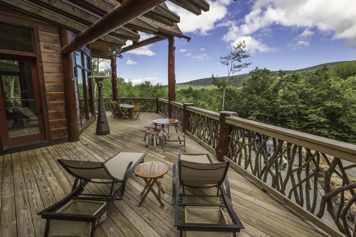 A spacious wooden deck furnished with lounge chairs and a dining table overlooks expansive mountain views. The scene captures the tranquil atmosphere of outdoor living, with the deck offering a seamless connection between the home and nature, ideal for relaxing, reading, and socializing while soaking up the vistas.