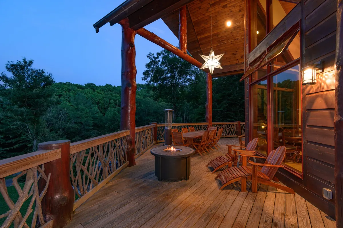 A spacious wooden deck features a central fire pit surrounded by rocking chairs and a star-shaped light fixture. The backdrop of towering trees and a tranquil seating area create an inviting spot for evening gatherings and stargazing.