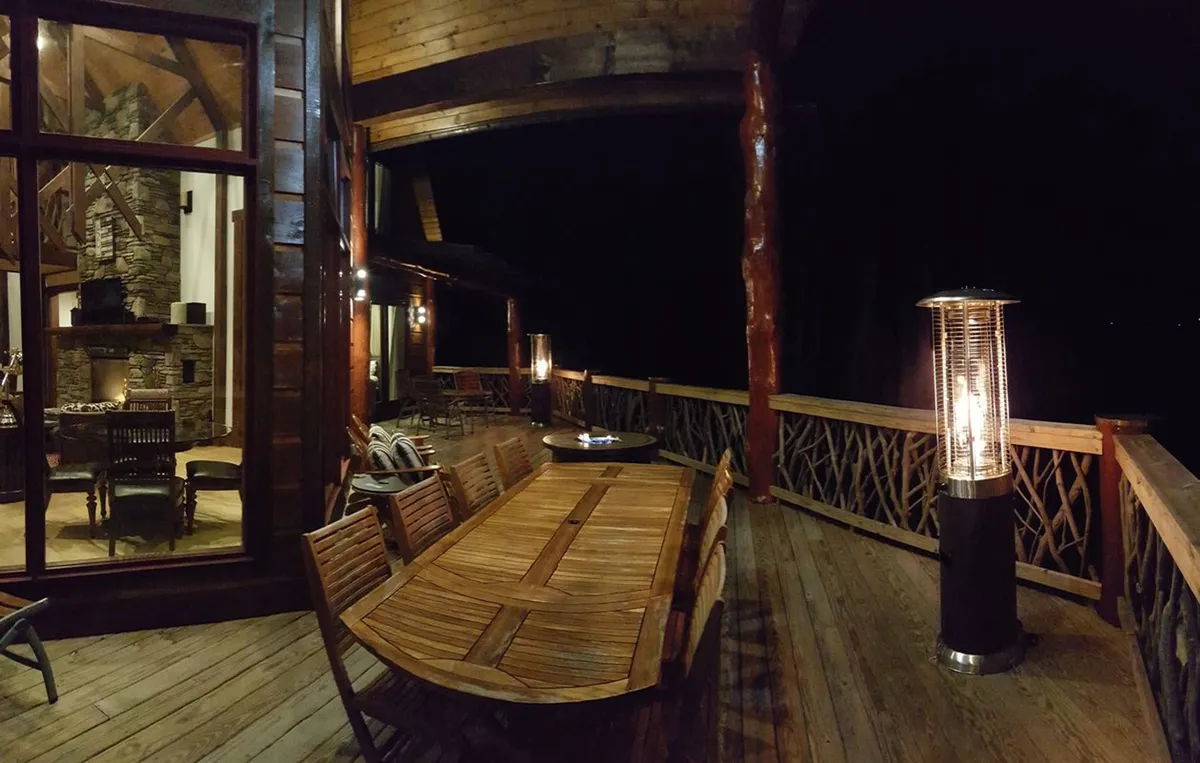 This exterior image features a beautifully set wooden dining table on a patio, complete with a heating lamp. The warm glow from the interior fireplace is visible through the large windows, creating a seamless connection between indoor comfort and outdoor enjoyment. The space invites gatherings under the open sky and emphasizes communal relaxation.