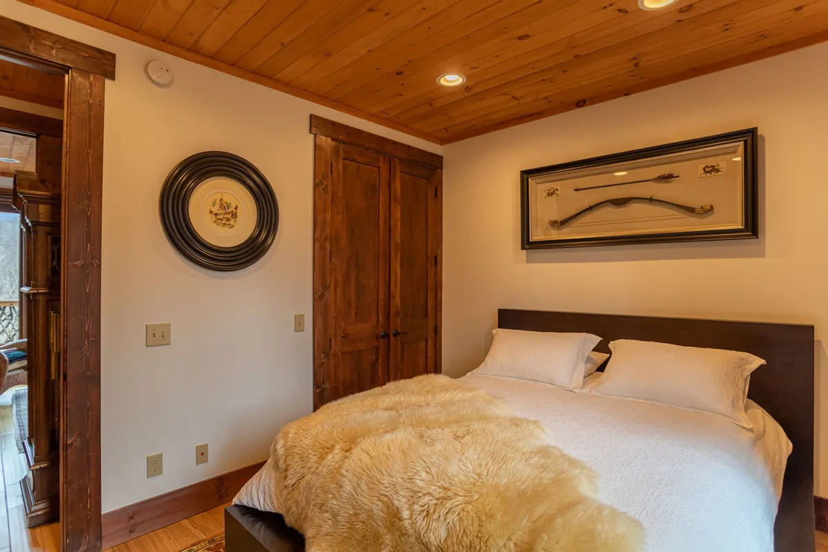 A cozy master bedroom featuring a plush bed with a fur throw, framed artwork on the walls, a warm wooden door, and inviting circular wall decoration. Natural light streams in, creating a tranquil and welcoming ambiance ideal for relaxation and restful sleep.