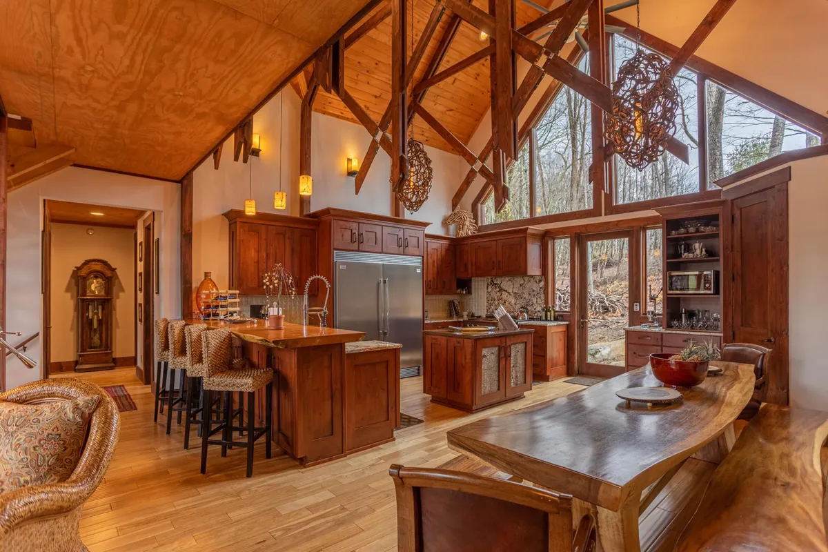 A view from the great room into the open kitchen and dining area, featuring a large wooden dining table, kitchen island, and exposed beams overhead. Generous windows bring in daylight and mountain views, underscoring the open-concept design. The setup is ideal for feasts, celebrations, and effortless flow between social spaces.