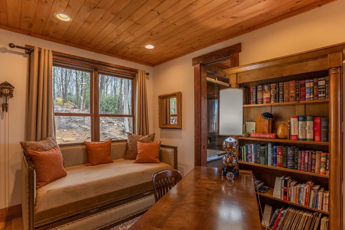 A peaceful library/loft area offering a daybed with plush cushions, a well-stocked bookshelf, a wooden desk, and a large window with a nature view. This tranquil multi-purpose space is ideal for reading, working, or extra sleeping arrangements for children.
