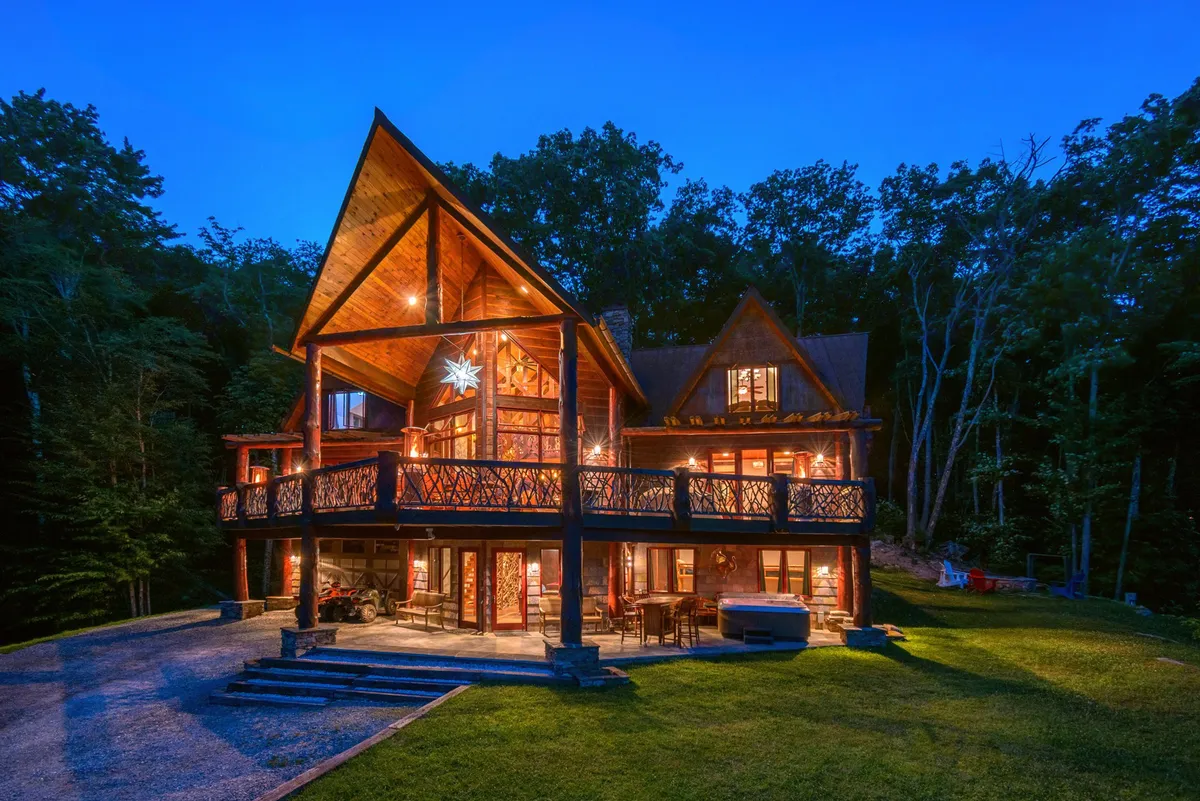 The hero image showcases the exterior of a custom mountain lodge at dusk, with the deck warmly lit and inviting. Architectural peaks create a striking silhouette against the sky, while surrounding trees add privacy and a serene, natural setting. Guests can envision relaxing in the hot tub, dining outdoors, or sipping wine on the deck as the sun sets over the Blue Ridge Mountains.