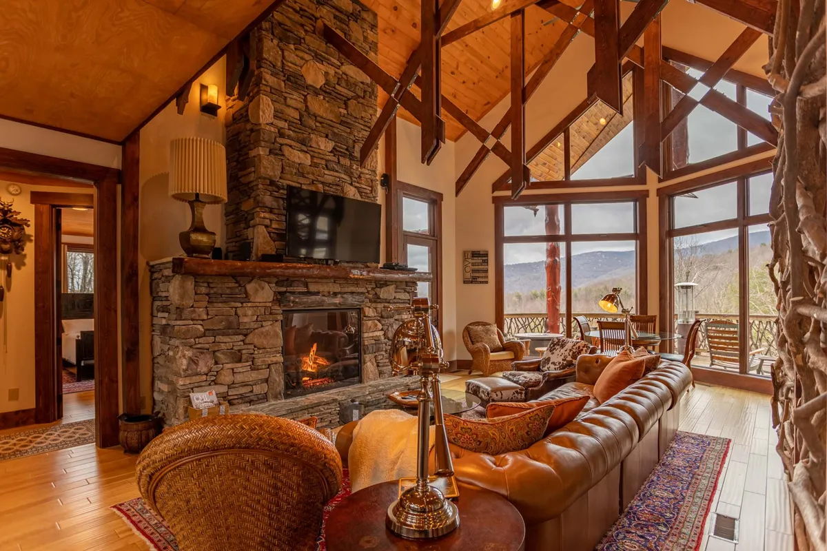 The image features a grand great room with a soaring ceiling accented by robust wooden beams. A substantial stone fireplace anchors one wall, while a deep leather sofa invites relaxation. Floor-to-ceiling windows showcase breathtaking mountain views, allowing natural light to flood the space and connect the interior to the outdoors. The open plan layout is visible, with sightlines to adjoining spaces, emphasizing the room’s role as a gathering hub.