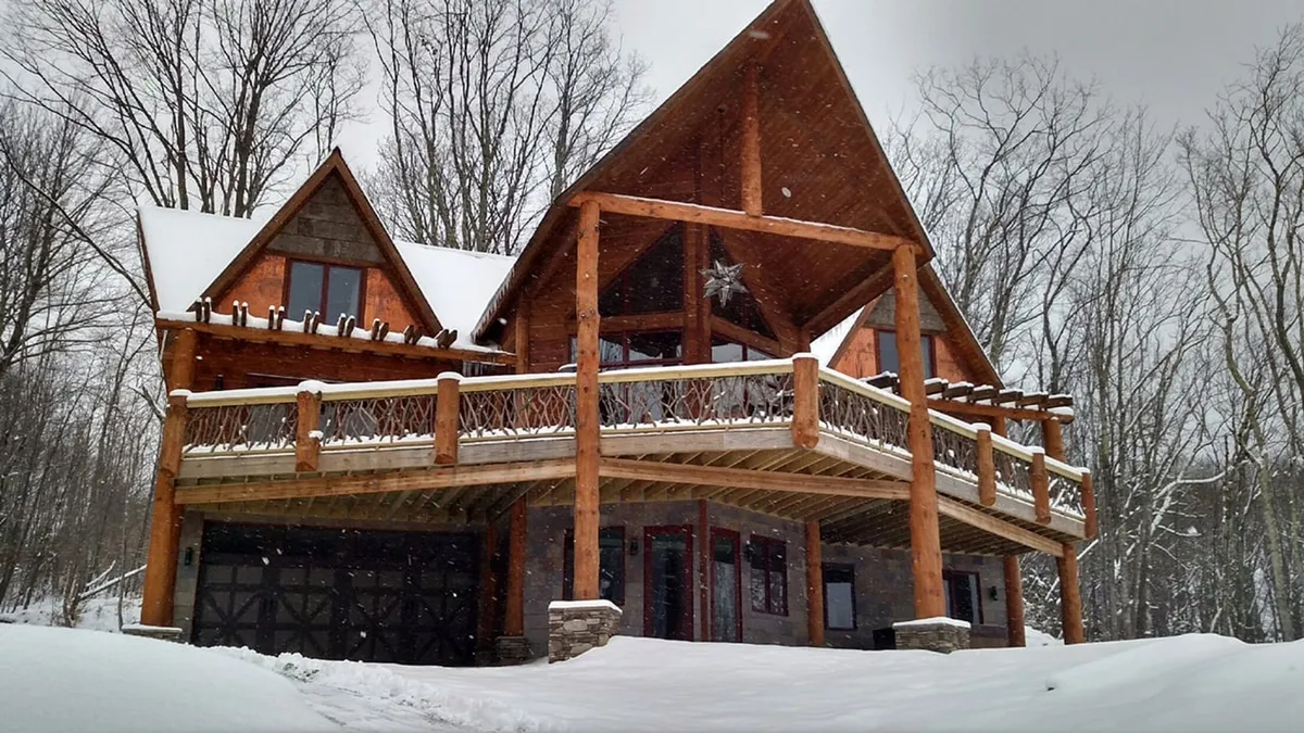 A large wooden deck framed by a triangular roof and stone accents, overlooking a snow-blanketed landscape. The scene suggests winter activities and gatherings, with footprints in the snow and the inviting glow from inside the home.