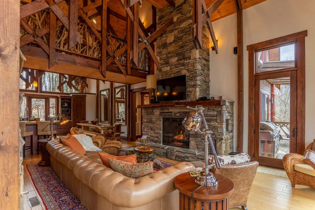 This inviting great room scene showcases a large, plush leather sectional sofa positioned around a substantial stone fireplace. The room features exposed wooden beams and expansive windows, providing views of the surrounding landscape and bathing the interior in natural light. The furniture arrangement encourages conversation and communal relaxation.