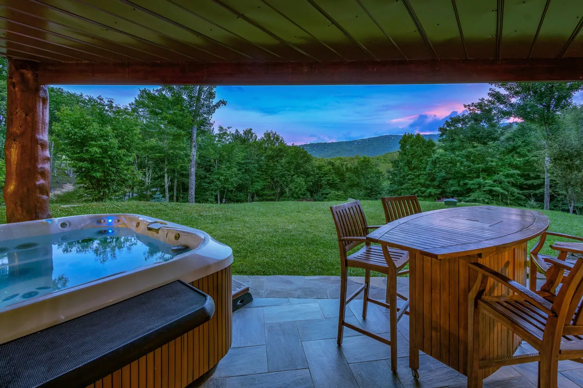 A tranquil deck area showcasing a hot tub and a dining table, set against a backdrop of sweeping mountain views. The scene is serene, ideal for relaxation, soaking in the hot tub, and dining with friends or family while taking in the natural surroundings.
