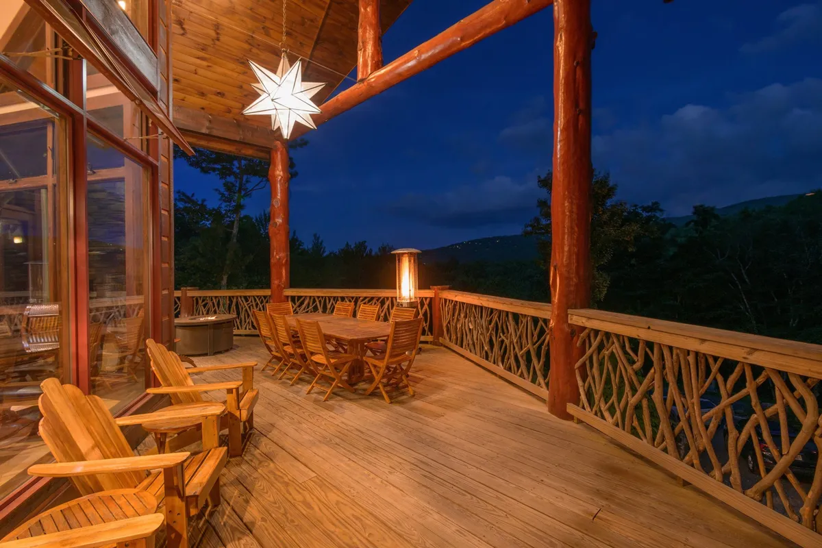 A large wooden deck with a dining table and wooden chairs positioned to face a panoramic mountain view. String lights and a star-shaped light fixture create a welcoming setting for social gatherings or quiet evenings under the stars.