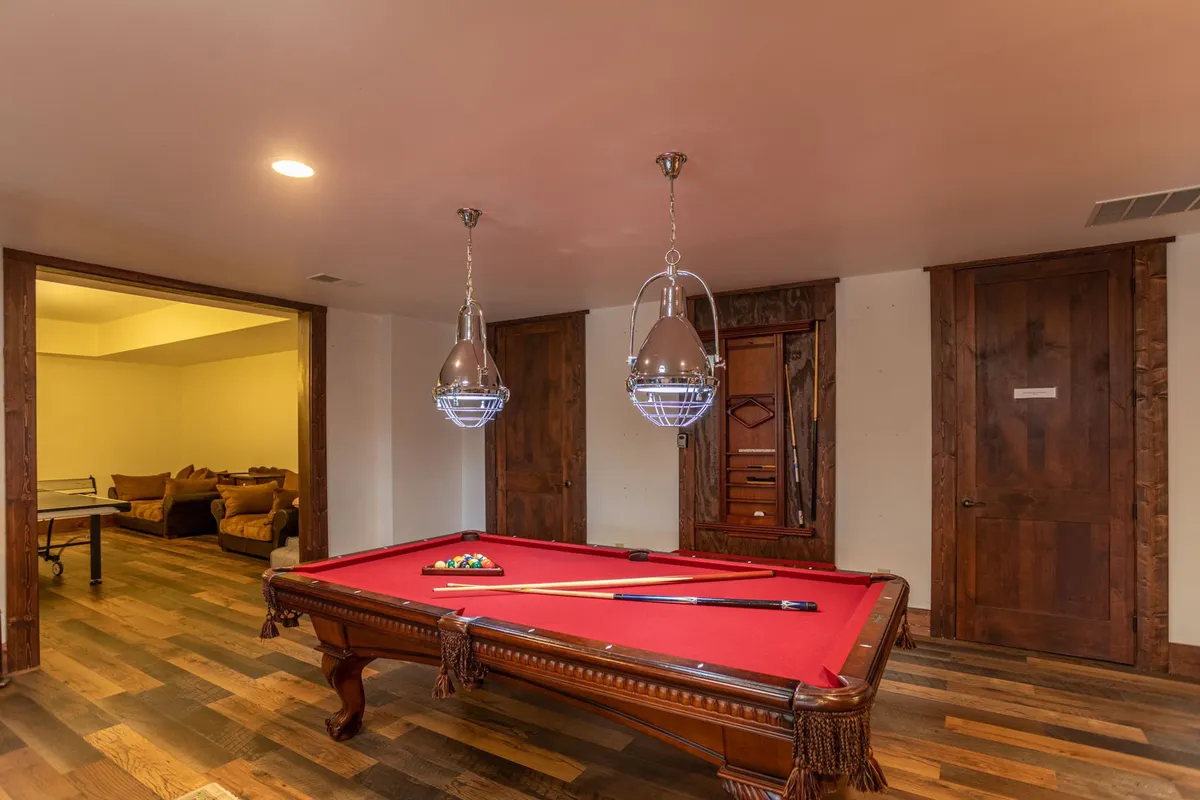 A professional pool table sits at the center of a stylish, inviting game room. Hanging light fixtures cast a warm glow, and wooden doors create a cozy, private atmosphere. The space encourages friendly competition and relaxed socializing, ideal for gatherings and game nights.